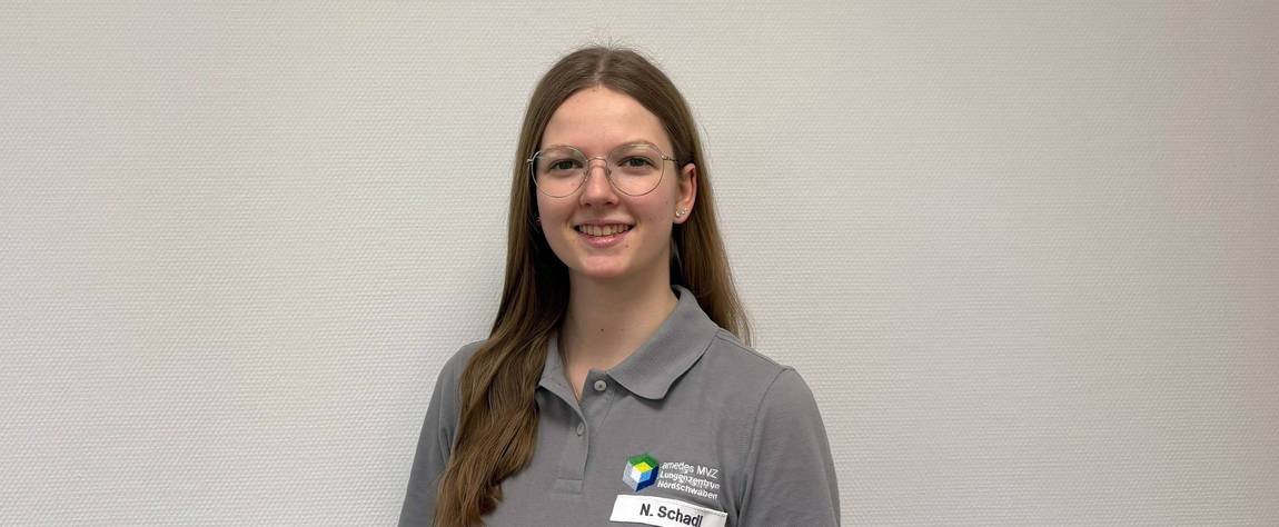 Eine junge Frau mit langen braunen Haaren und Brille lächelt in die Kamera. Sie trägt ein graues Poloshirt mit einem Logo und einem Namensschild mit der Aufschrift N. Schadl und steht vor einer schlichten hellen Wand.