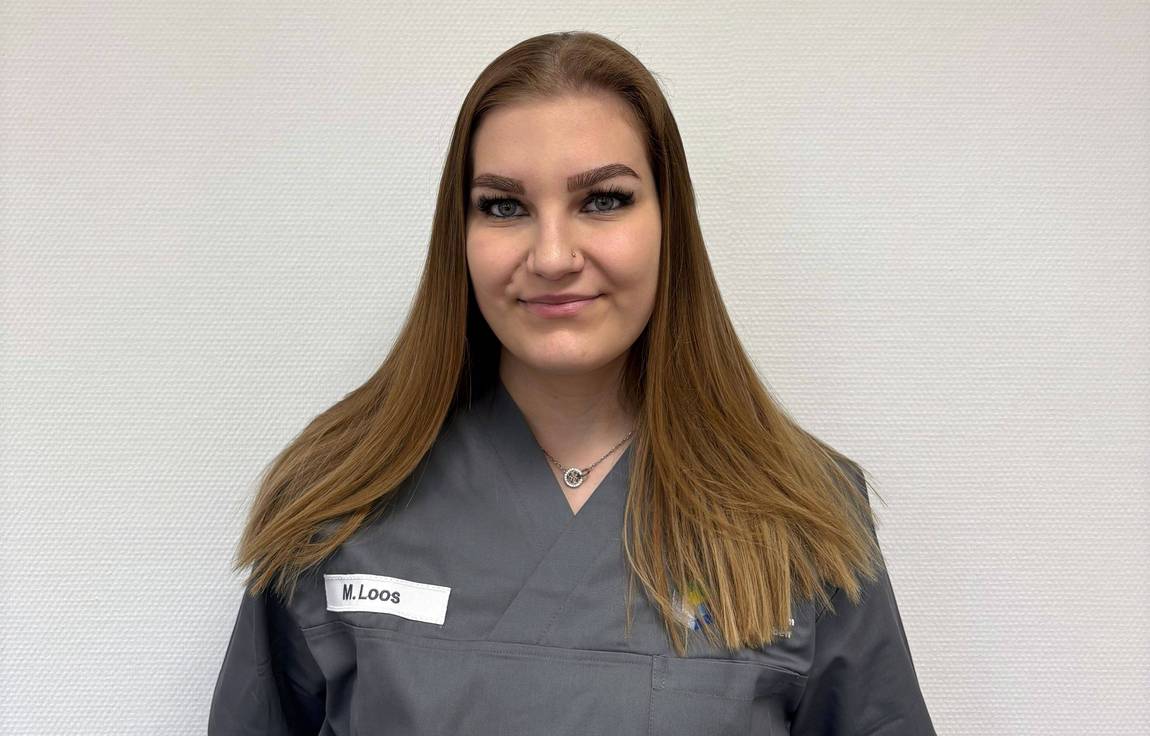 Eine Frau mit langen hellbraunen Haaren, die einen grauen Arztkittel trägt, steht vor einer schlichten weißen Wand. Sie trägt ein Namensschild mit der Aufschrift M. Loos und lächelt sanft in die Kamera.