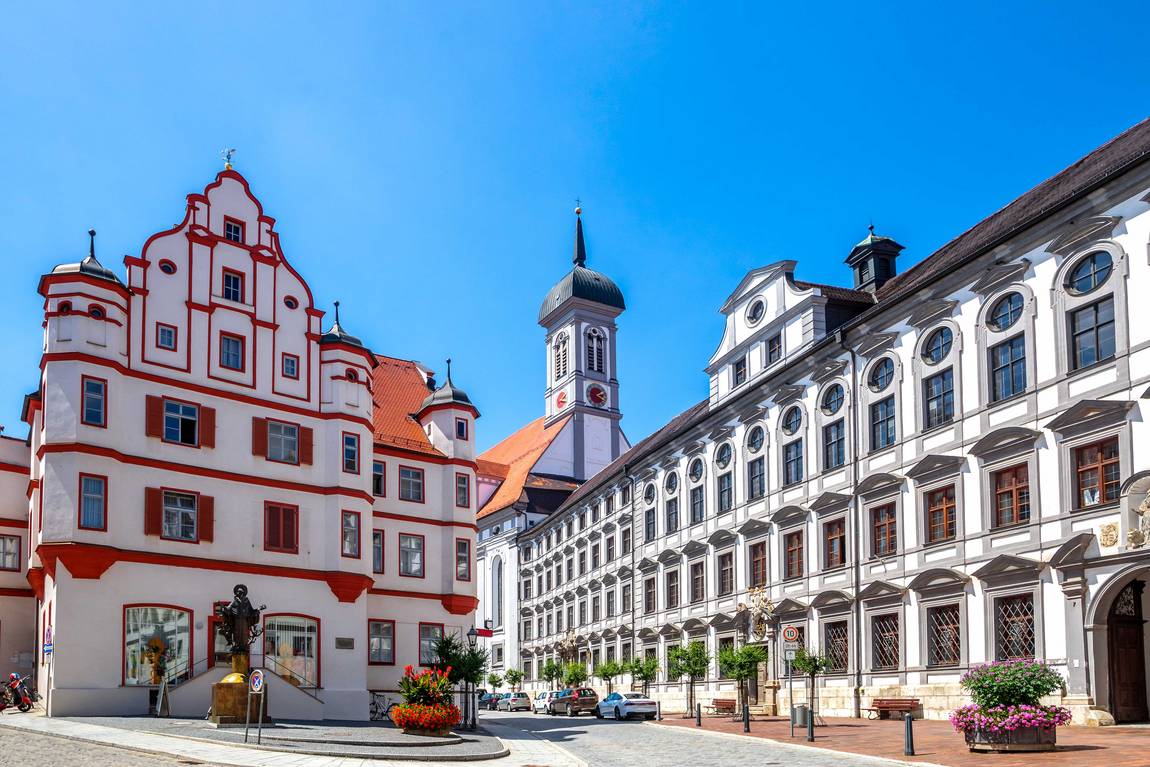 Eine sonnige Straße in Augsburg, Deutschland, mit dem historischen weiß-roten Perlachturm, verschnörkelten Barockgebäuden, Blumenkübeln und einem strahlend blauen Himmel über der Stadt.