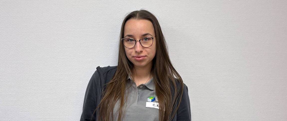 Eine junge Frau mit langen braunen Haaren und Brille steht vor einer weißen, strukturierten Wand. Sie trägt ein graues Poloshirt, eine dunkle Jacke und ein Namensschild. Sie blickt mit neutralem Blick in die Kamera.