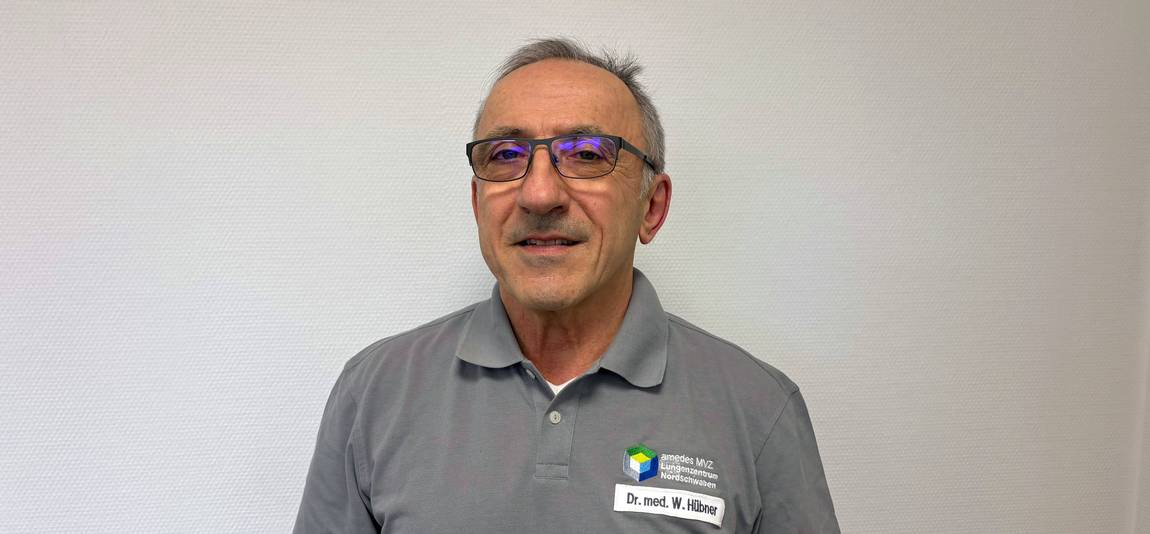 Ein Mann mit kurzen grauen Haaren und Brille, der ein graues Polohemd mit einem Namensschild trägt, auf dem Dr. med. W. Hübner steht. W. Hübner steht vor einer schlichten weißen Wand.