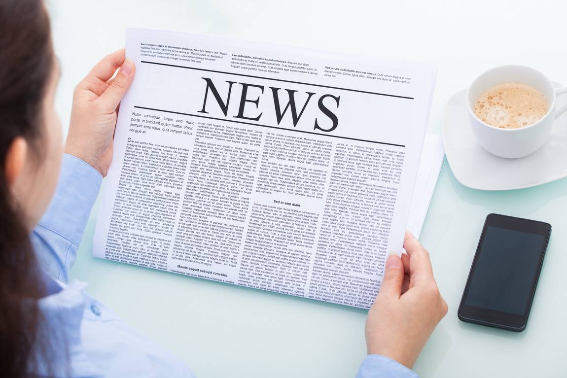 Eine Person hält eine Zeitung mit der Schlagzeile NEWS in der Hand und liest sie an einem Tisch. In der Nähe befinden sich ein Smartphone und eine Tasse Kaffee. Die Person ist von hinten zu sehen.