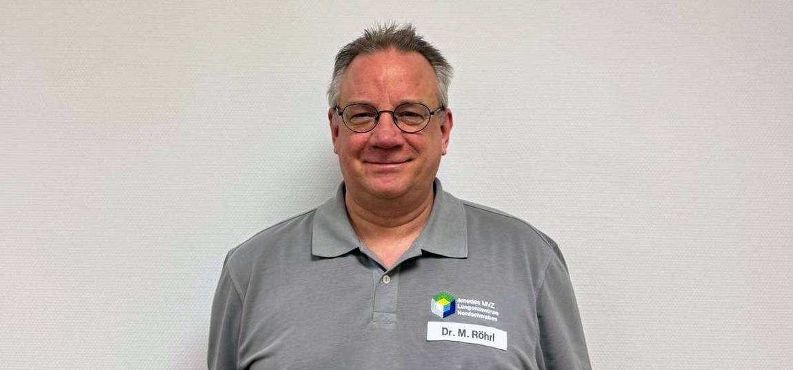 Ein Mann mit Brille und kurzen grauen Haaren trägt ein graues Poloshirt mit einem Firmenlogo und einem Namensschild mit der Aufschrift Dr. M. Röhl. Er steht vor einer schlichten hellen Wand und lächelt leicht.