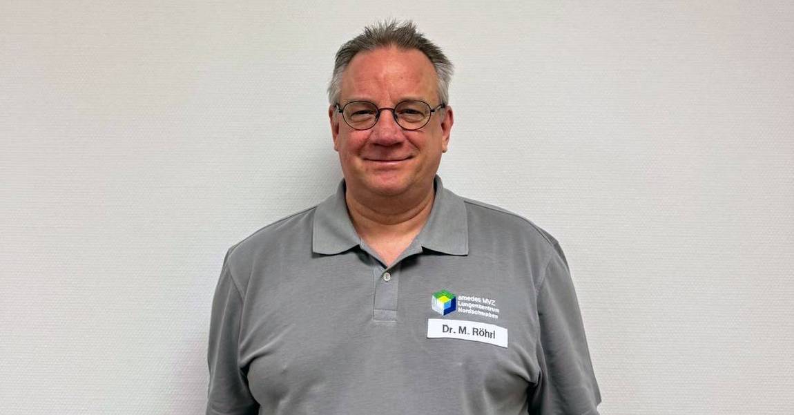 Ein Mann mit Brille und kurzen grauen Haaren trägt ein graues Poloshirt mit einem Firmenlogo und einem Namensschild mit der Aufschrift Dr. M. Röhl. Er steht vor einer schlichten hellen Wand und lächelt leicht.