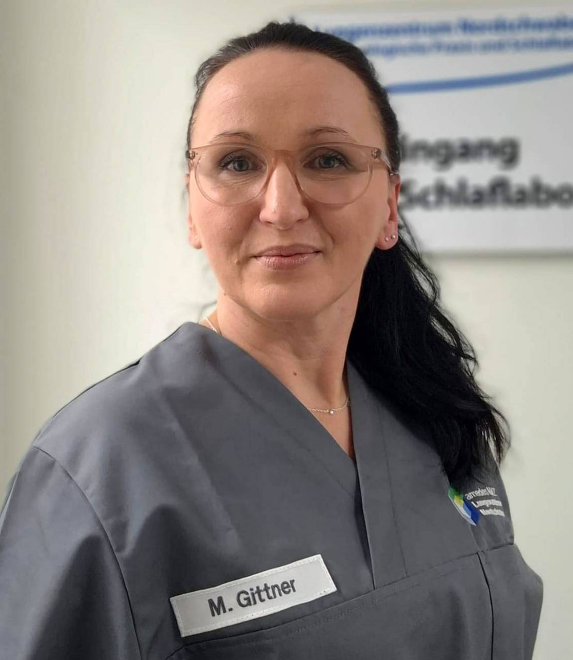Eine Frau mit Brille, die einen grauen Arztkittel und ein Namensschild mit der Aufschrift M. Gittner trägt, steht in einem Raum mit einem verschwommenen Schild im Hintergrund.