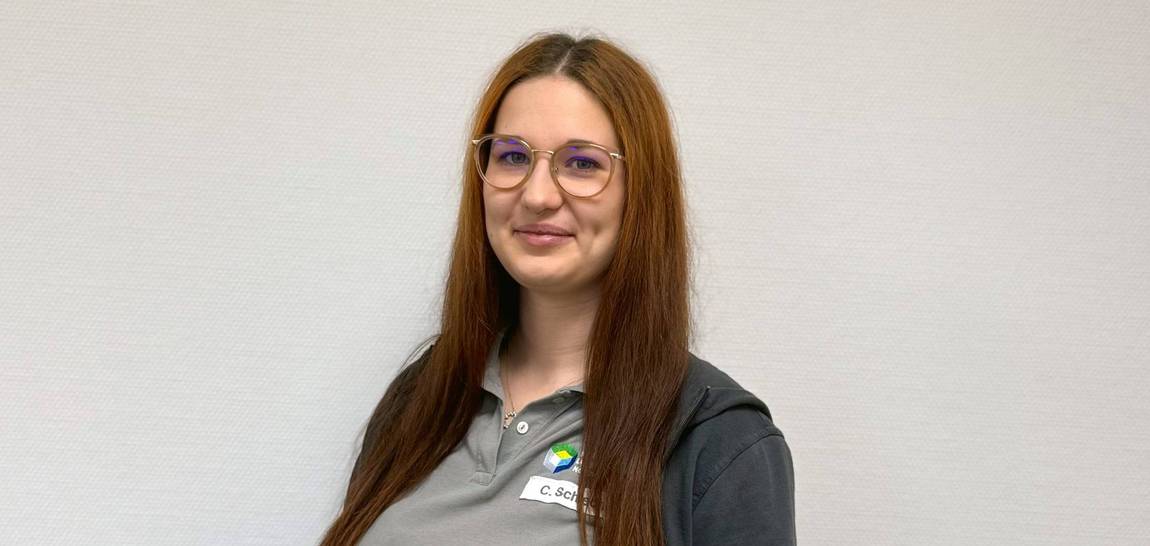 Eine Frau mit langen braunen Haaren und Brille, die ein graues Poloshirt mit Logo und eine schwarze Jacke trägt, steht lächelnd vor einer hellen Wand.
