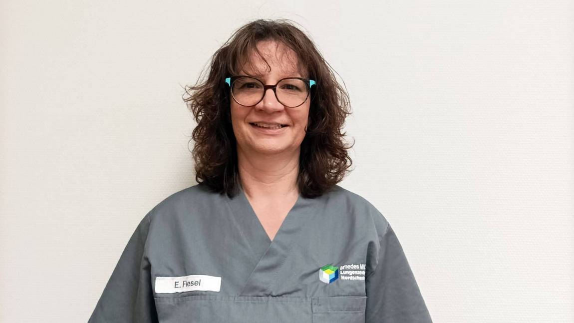 Eine Frau mit Brille und grauem Arztkittel steht lächelnd vor einer schlichten weißen Wand. Auf dem Namensschild an ihrem Kittel steht E. Füssel.