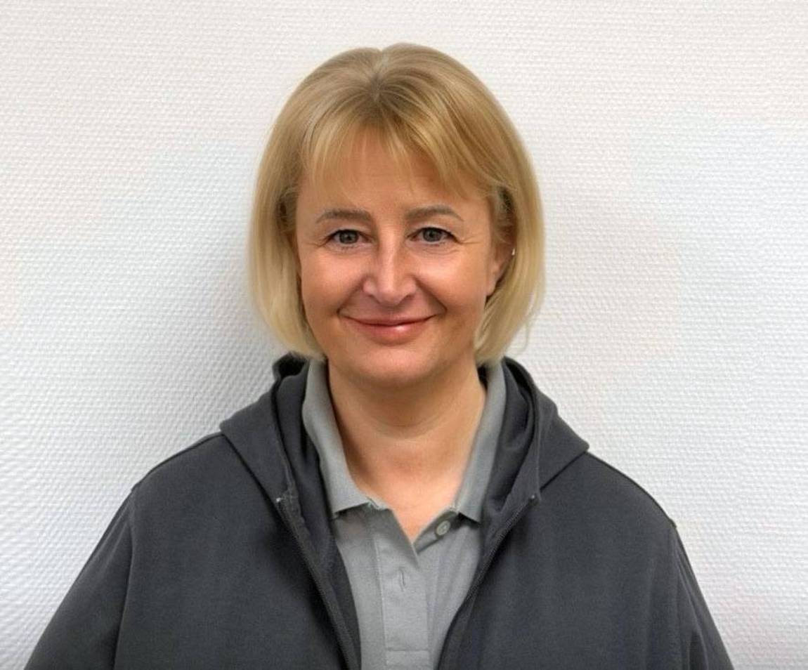 Eine Frau mit kurzen blonden Haaren, die ein graues Hemd mit Kragen und eine graue Jacke mit Reißverschluss trägt, steht lächelnd vor einer schlichten weißen, strukturierten Wand.