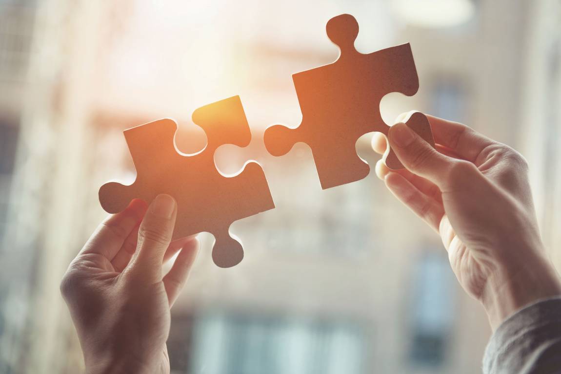 Zwei Hände halten große Puzzlestücke aufeinander zu und richten sie mit dem durchscheinenden Sonnenlicht aus, das Verbindung oder Problemlösung symbolisiert, mit einem unscharfen Innenhintergrund.
