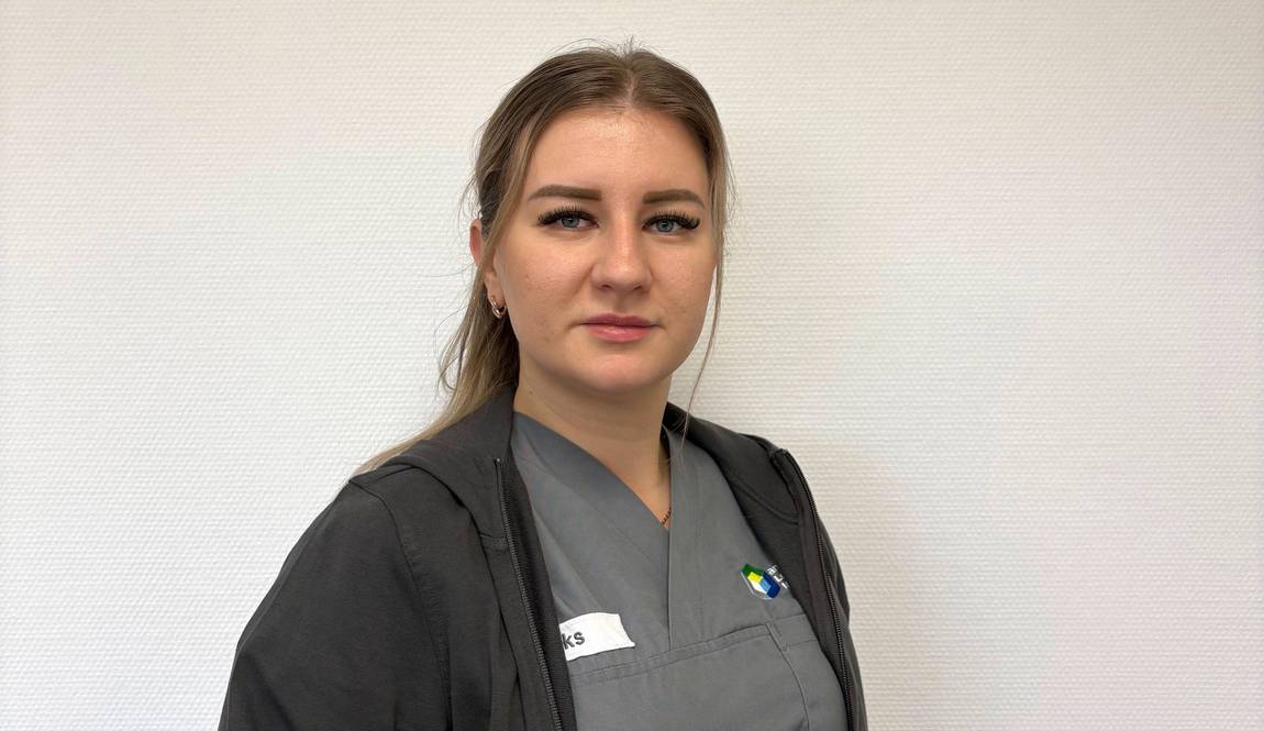 Eine Frau in grauem Arztkittel und schwarzem Kapuzenpulli steht vor einer schlichten weißen Wand und schaut mit neutralem Blick in die Kamera. Sie hat ihr hellbraunes Haar zurückgebunden und trägt ein Namensschild auf der Brust.