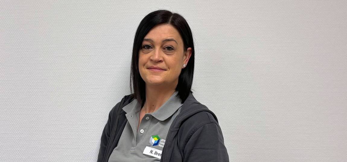 Eine Frau mit glattem dunklem Haar steht vor einer hellen, strukturierten Wand und trägt eine graue Jacke mit Reißverschluss über einem grauen Poloshirt mit Namensschild. Sie blickt in die Kamera und lächelt leicht.