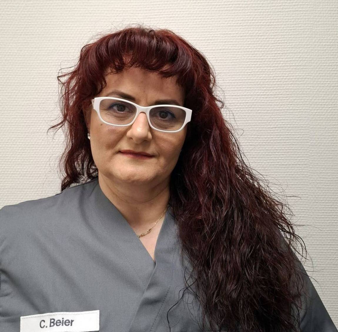 Eine Frau mit langen, gewellten dunkelroten Haaren, einer Brille und einer grauen Uniform mit einem Namensschild, auf dem C. Beier steht, steht vor einer hellen, strukturierten Wand.