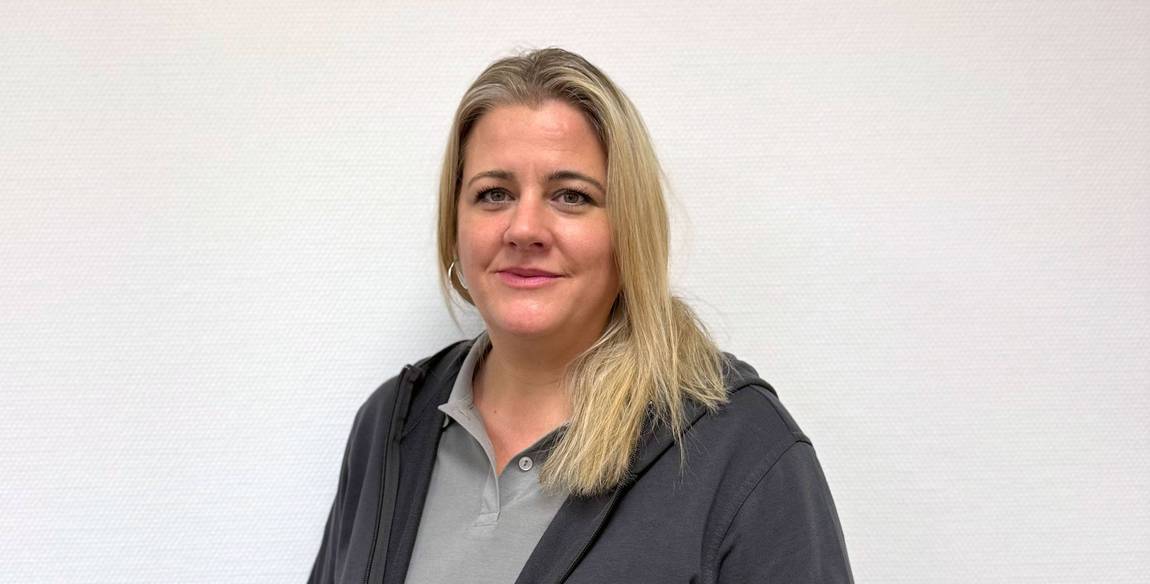 Eine Frau mit langen blonden Haaren, die einen grauen Kapuzenpullover mit Reißverschluss über einem hellgrauen Poloshirt trägt, steht vor einem schlichten weißen Hintergrund und lächelt leicht in die Kamera.