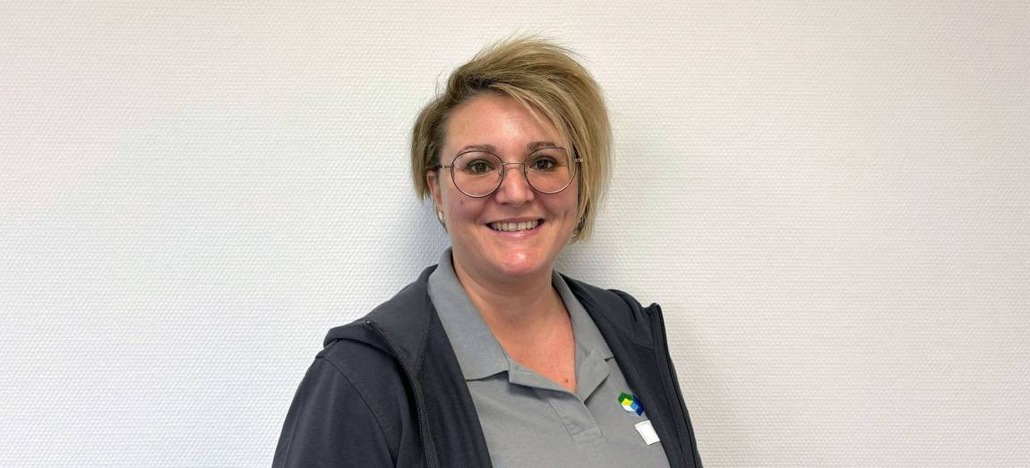 Eine lächelnde Person mit kurzen, hellen Haaren und Brille, die ein graues Poloshirt und eine dunkle Jacke mit Reißverschluss trägt, steht vor einer schlichten, hellen Wand.