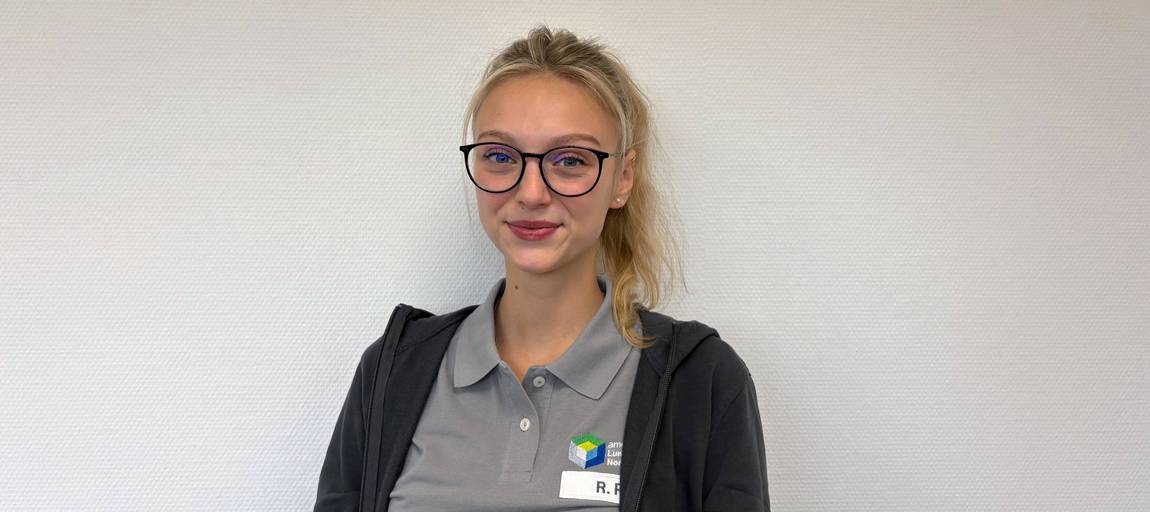 Eine Frau mit langen blonden Haaren in einem Pferdeschwanz, die eine Brille, ein graues Polohemd mit Logo, einen Kapuzenpulli und ein Namensschild trägt, steht vor einer schlichten weißen Wand und lächelt leicht.