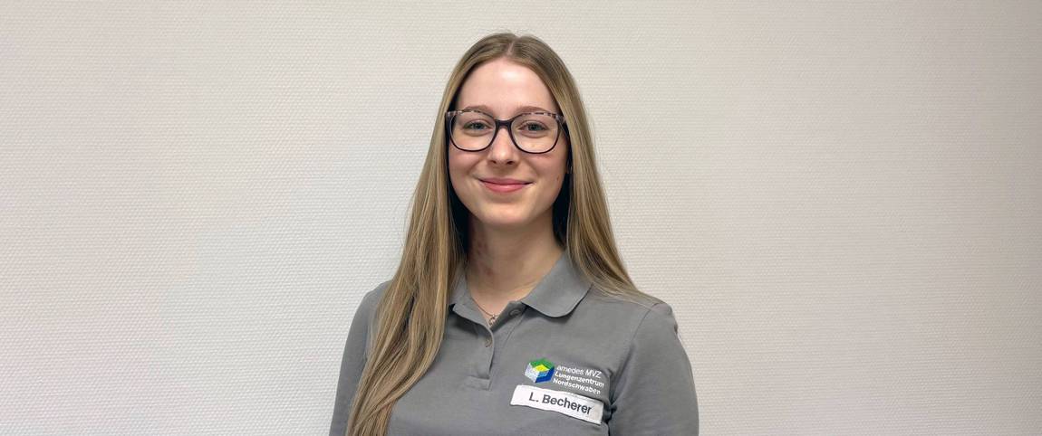 Eine junge Frau mit langen blonden Haaren, Brille und einem grauen Poloshirt mit Firmenlogo und einem Namensschild, auf dem L. Bechter steht, steht lächelnd vor einer schlichten hellen Wand.