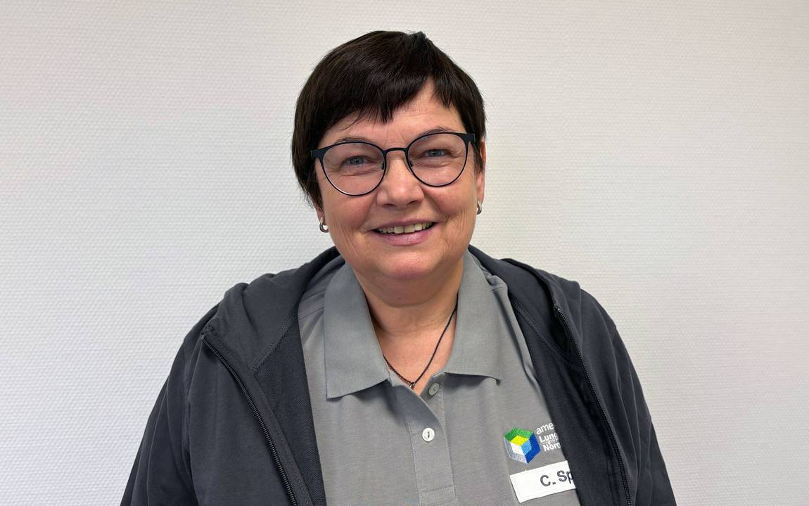 Eine Person mit kurzen dunklen Haaren und Brille lächelt in die Kamera. Sie trägt ein graues Hemd mit Kragen, eine dunkle Jacke und ein Namensschild mit der Aufschrift C. S. und einem bunten Logo. Der Hintergrund ist eine schlichte helle Wand.